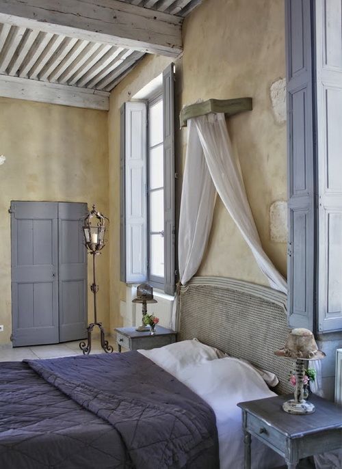 a Provence bedroom with tan walls, a vintage bed with a cane headboard, blue and white bedding, blue furniture, shutters and textiles