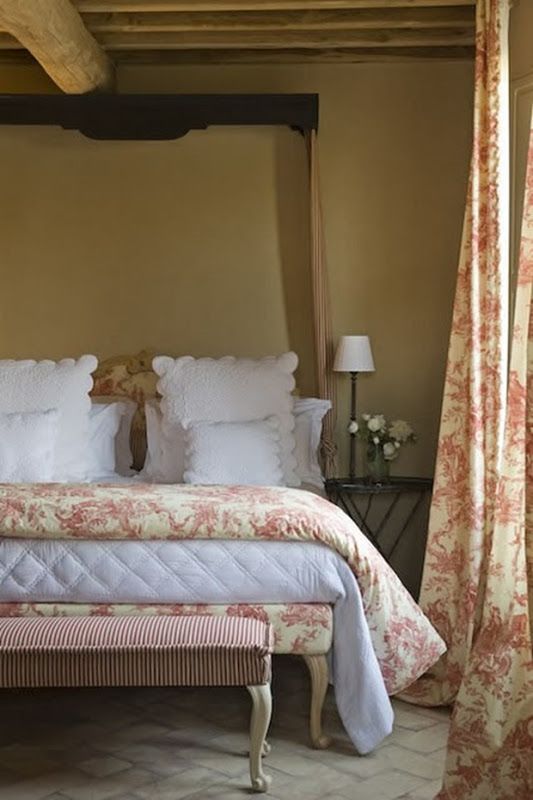 a Provence bedroom with tan walls, a wooden ceiling with wooden beams, a bed with pastel bedding, a refined bench and floral curtains