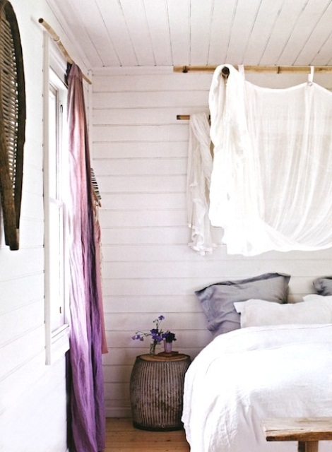 a shabby chic and rustic feminine bedroom in neutrals accented with lavender pillows, purple curtains and a neutral semi sheer canopy over the bed