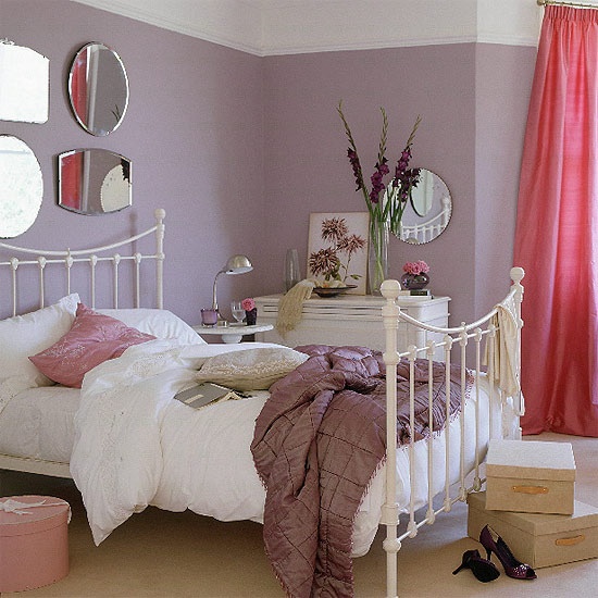 a feminine bedroom with lilac walls, a white forged bed, pink curtains, a gallery wall of mirrors and some artworks and blooms