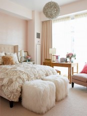 a pretty pink bedroom with a neutral bed, white fur stools, a simple table and a chair, a floral chandelier and pink curtains