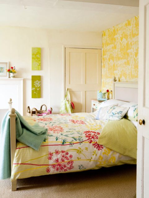 a bright feminine bedroom with an accent yellow botanical wall, neutral furniture and a faux fireplace, bright floral bedding and bold artworks