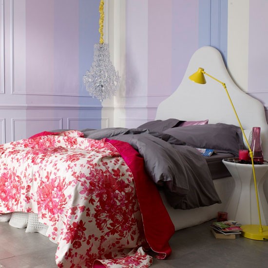a delicate bedroom done in pastels, with striped walls, a creamy bed and a nightstand, purple and red printed bedding and gold touches for romantic people