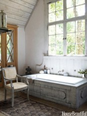 a chic neutral barn bathroom with wooden planks on the walls, a wood clad bathtub, vintage furniture and a large window to flood the space with light