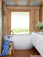 a modern barn bathroom clad with wood, with wooden beams, an oval tub and a sleek white vanity plus a large window