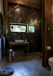 a dark barn bathroom clad with wood inspried tiles, with wooden beams, a concrete floor and a mirror in a metal frame