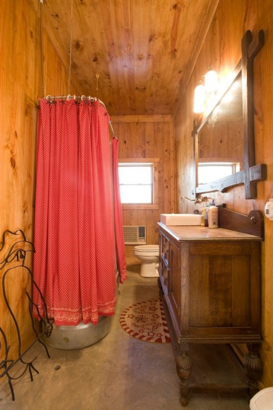 a warm barn bathroom clad with wood, a wooden vanity, a large mirror, a metal tub and a bright red curtain