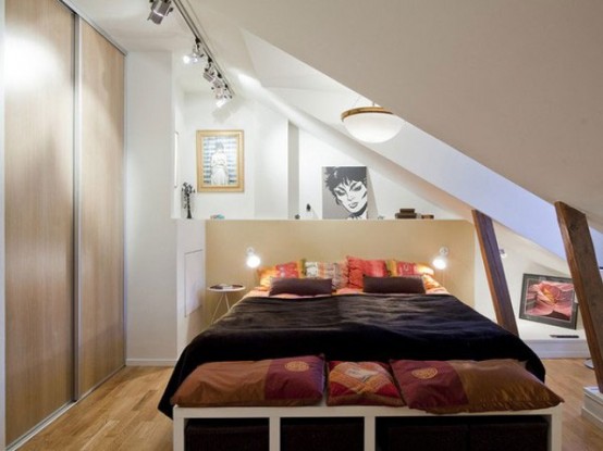 a tiny attic bedroom with wooden beams, a large window, a bed with lights and dark textiles plus a storage unit