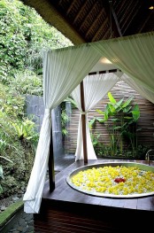 a sunken outdoor bathtub in a wooden platform with curtains for privacy, an outdoor shower and some greenery planted