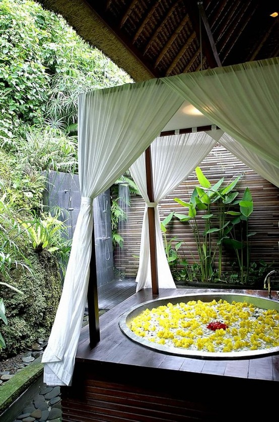 a sunken outdoor bathtub in a wooden platform with curtains for privacy, an outdoor shower and some greenery planted