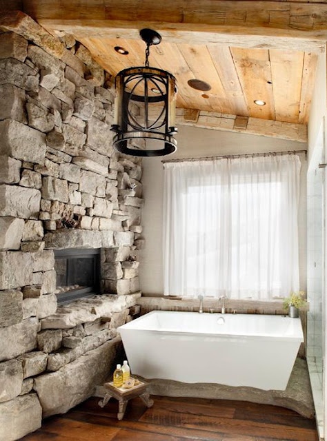 a farmhouse or cabin bathroom with a stone fireplace, a rectangular bathtub, neutral curtains and a metal pendant lamp