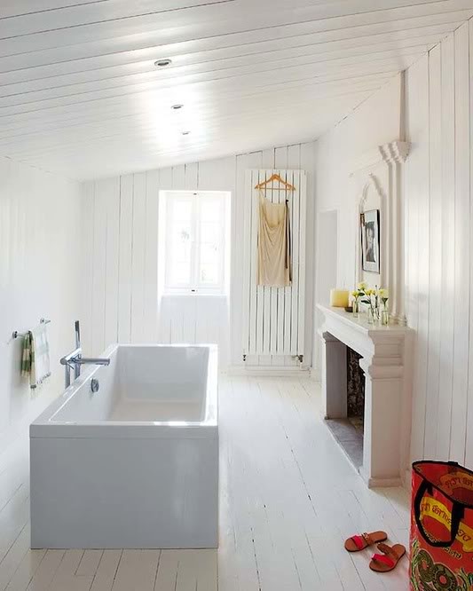 a chic white bathroom clad with planks, a rectangular bathtub, a fireplace with a mantel and some decor and a radiator on the wall