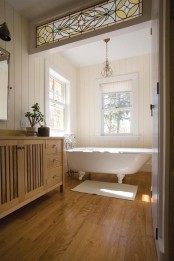 a neutral vintage bathroom with a clawfoot bathtub, a stained vanity, a wooden floor, windows and a beautiful stained glass detail that brings interest to the space