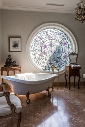 a fantastic and jaw-dropping vintage-inspired bathroom with a round window with stained glass, a clawfoot bathtub and vintage carved furniture just wows
