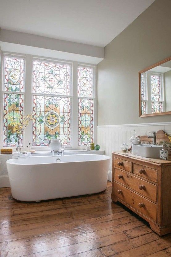an airy farmhouse bathroom with olive green walls, a wooden floor, a stained vanity, an oval tub and a large window with stained glass that keeps privacy of the space