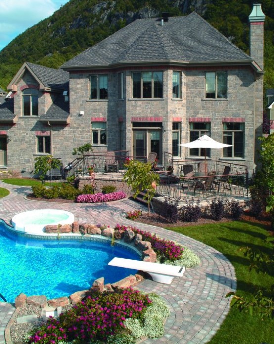 a stone house with a stone pool deck, a built-in jacuzzi and a pool, with bold blooms and greenery and green lawn to refresh the look