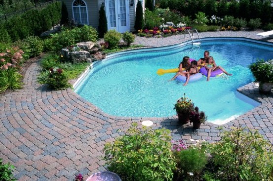 a catchily shaped pool with a stone deck, lots of greenery and blooms around that refresh the stone deck look compose a lovely and cozy space to be in
