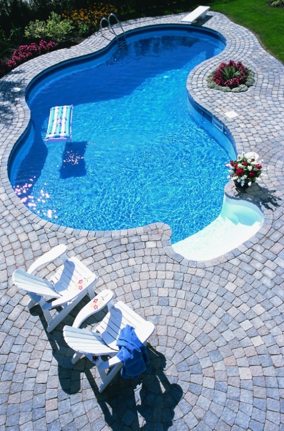 a catchily shaped pool with a whitewashed stone deck, simple white outdoor furniture, green lawn and bright blooms