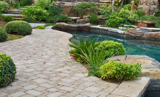 a pretty backyard with a catchily shaped pool, a neutral stone deck, planted greenery and some blooms is a lovely outdoor space
