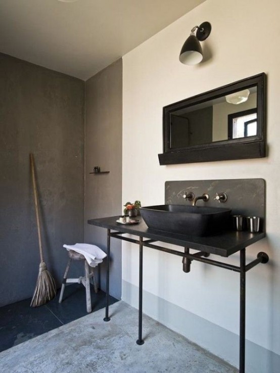 a modern industrial bathroom with a concrete floor and walls, a black sink on a vanity, a mirror and a wall sconce