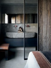a modern industrial bathroom with concrete and rough wood walls, a large carved stone sink, a long mirror and a leather bench