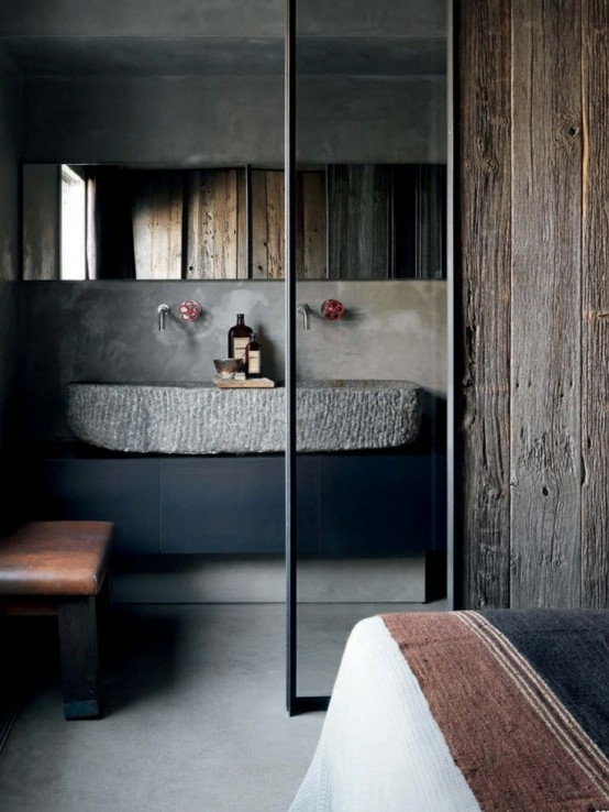 a modern industrial bathroom with concrete and rough wood walls, a large carved stone sink, a long mirror and a leather bench