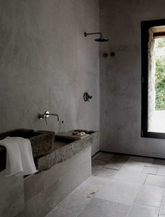 a minimalist industrial bathroom done with concrete and tiles, stone sinks, exposed pipes and a large window for a view