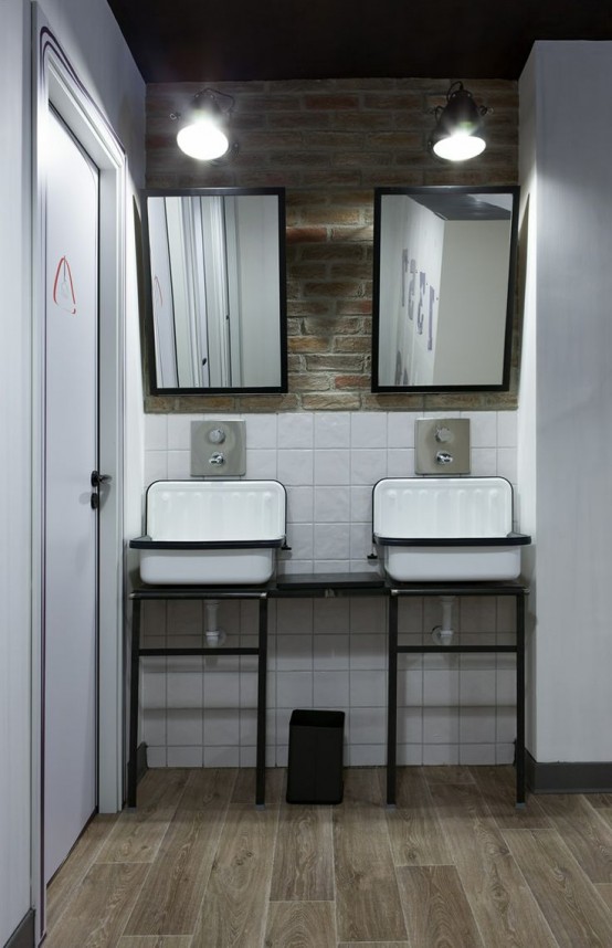 a modern industrial bathroom with white tiles, a brick wall, a wooden floor, vintage sinks on a stand and a duo of mirrors