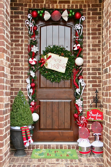 a bright and fun Christmas front door with a topiary with striped legs, candies in buckets, an evergreen wreath and a colorful garland with ornaments and toy candies