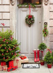 a Christmas front door with a festive greenery wreath with a bow, a posie and a large Christmas tree with red ornaments and mini trees in pots