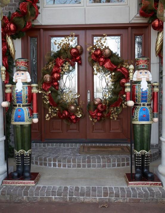 whimsical Christmas front door styling with evergreens, ornaments, gilded leaves and red ribbons plus giant nutcrackers on front of the door