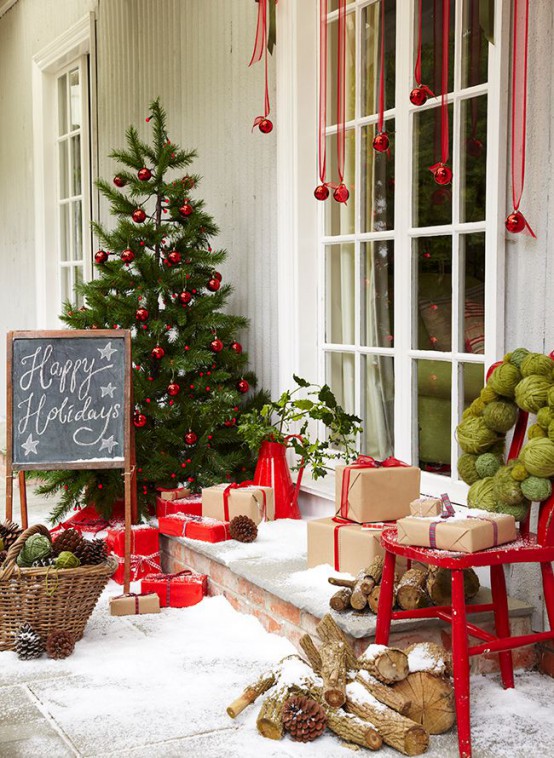 a red and green Christmas front door styled with a Christmas tree with red ornaments, red ornaments hanging down, gift boxes, firewood and a basket with pinecones and yarn balls