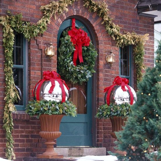 a Christmas front door with an oversized greenery wreath with a red bow, two urns with evergreens and oversized silver ornaments and red bows looks cool