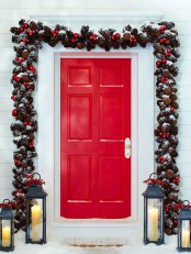 bold Christmas front door styling with a garland made of pinecones and red ornaments that frames the door and candle lanterns