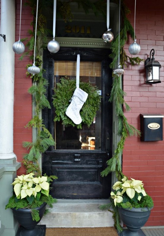 natural Christmas front door decor with hanging silver ornaments, an evergreen garland and wreath with a stocking and an urn with neutral blooms