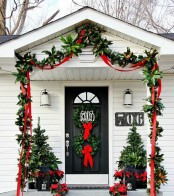 a cozy holiday front door with an evergreen garland, a duo of wreaths and mini Christmas trees plus fabric blooms and red bows