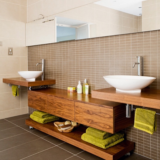 a modern bathroom clad with tan and brown tiles of various sizes, rich stained wooden vanities and a storage unit, bright towels