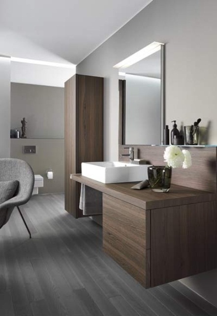 a minimalist bathroom with a grye laminate floor, stained wooden cabinets and a comfy grey chair plus a mirror