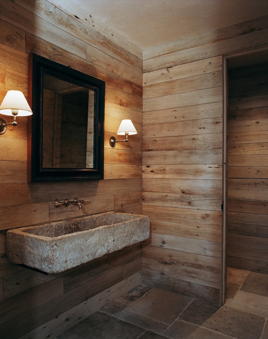 a rustic bathroom clad with light stained wood and with tiles on the floor, with a wall-mounted stone sink and sconces