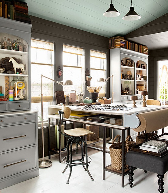 an eclectic home office done in neutrals, a tall desk, bookcases and a black ottoman