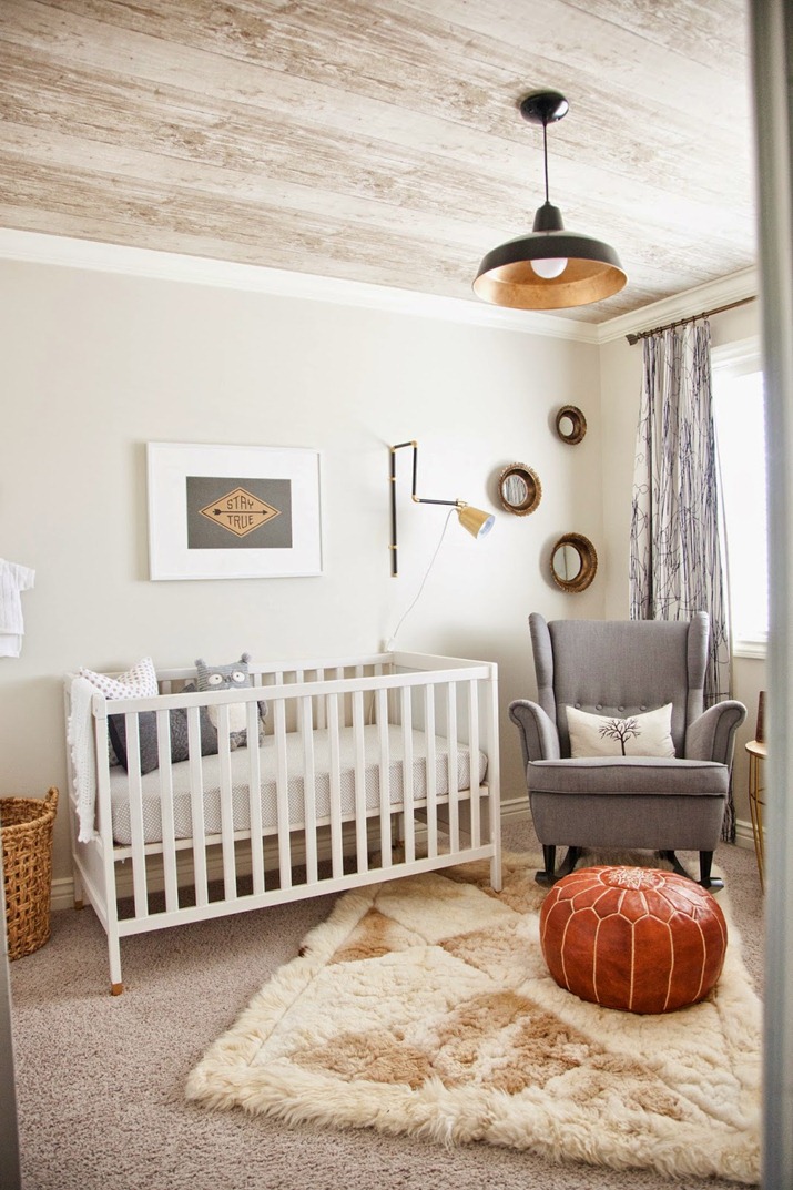 a neutral rustic inspired nursery with tan walls, a wood grain ceiling, layered rugs, a grey rocker chair and a leather pouf