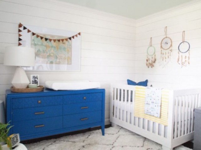 a welcoming farmhouse nursery with a bold blue dresser that is a changing table, a crib with blue and yellow bedding, dream catchers and a map as an artwork