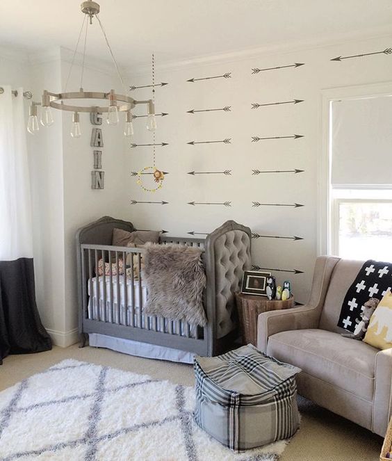 an eclectic nursery with an arrow accent wall, a grey crib with a tufted detail, a taupe chair and printed textiles, a pouf is a chic space