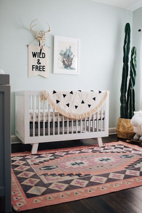a boho desert nursery with light grey walls, a white crib with printed bedding and a rug, a potted cactus and a mini gallery wall