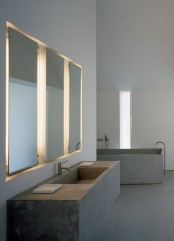 an ultra-minimalist concrete bathroom with a concrete bathtub and a floating vanity with a built-in sink plus mirrors with built-in lights