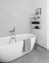 a white minimalist bathroom with a white tub, some wall shelves and a grey concrete floor