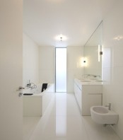 a white minimalist bathroom with large scale white tiles all over, a long double vanity and white appliances plus a vertical window with frosted glass