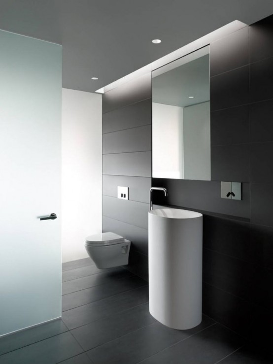 a dark minimalist powder room clad with long narrow tiles, a hidden storage unit and a frosted glass door