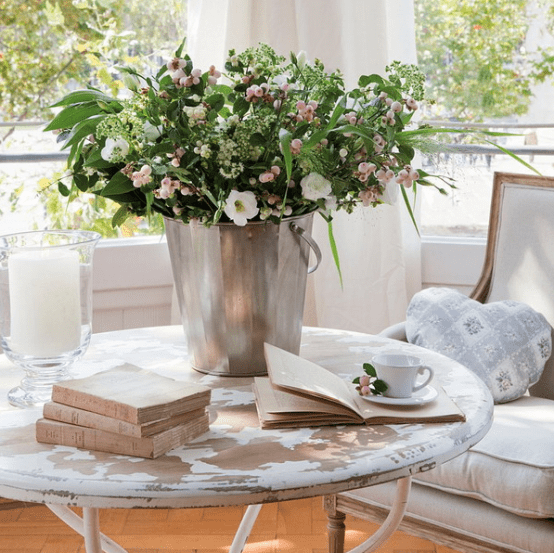 a round shabby chic coffee table with books, an oversized candle in a clear glass vase, a metal bucket with greenery and blooms