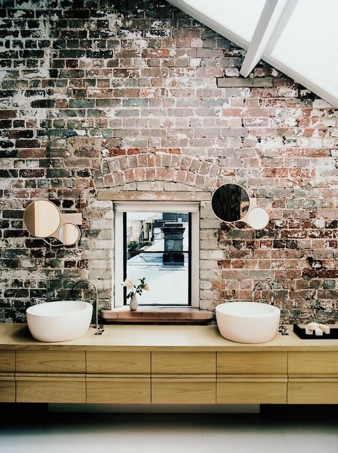 a modern bathroom with a red brick wall, a window, a long vanity with two sinks and a frosted glass roof